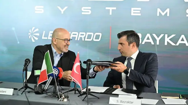 Leonardo CEO'su Roberto Cingolani ve Baykar yöneticisi Selçuk Bayraktar birbirlerine bakarak birbirlerine belge veriyor. Önlerinde İtalya ve Türkiye bayrakları var