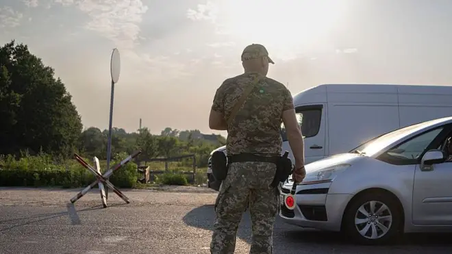 Чоловік у військовому камуфляжі стоїть спиною до камери посеред дороги біля імпровізованого блокпоста. Праворуч зупинений легковий автомобіль, у вікні якого видно водія. Позаду військовослужбовця – білий фургон. На узбіччі – дерев’яний шлагбаум, а вдалині – зелені дерева та відкрита місцевість.