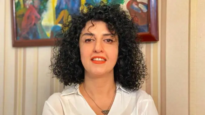Narges Mohammadi sonríe en esta foto de folleto sin fecha. Tiene el pelo negro rizado y lleva una camisa blanca, un collar en forma de triángulo y lápiz labial rojo.