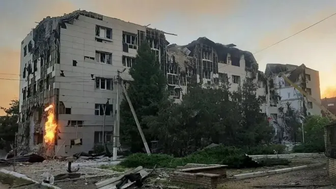 Duży, szary, masywny budynek jest wyraźnie uszkodzony i otoczony gruzem oraz rumowiskiem. Po jednej stronie budynku widoczne są płomienie.
