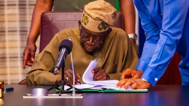 Aarẹ Bola Tinubu lasiko to n buwọ lu iwe nileeṣẹ Aarẹ, o mu gege dani, bẹẹ ni ọwọ kan wa lori tabili rẹ, nigba ti ẹnikan duro lẹyin rẹ.