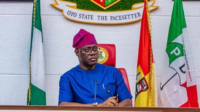 Govnor Seyi Makinde wearing blue clothes wit cap sitting between one Nigeria flag and PDP flag