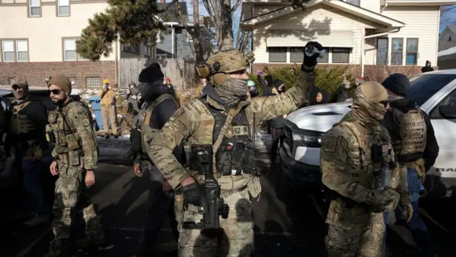 一軒家が並ぶ住宅街に、重装備で顔を隠した迷彩服姿の当局者が複数立っている