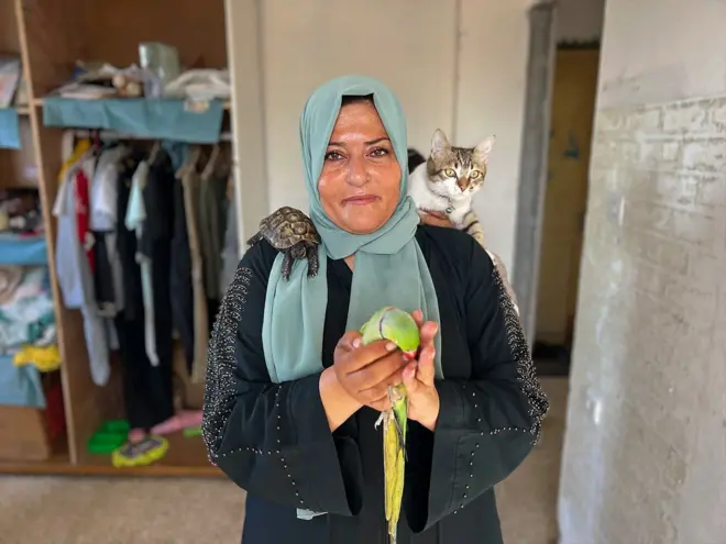 青系のスカーフを頭に巻き、黒い服を着たパレスチナ人女性が、緑色のインコを両手で持っている。左肩にはネコが、右肩にはカメが乗っている