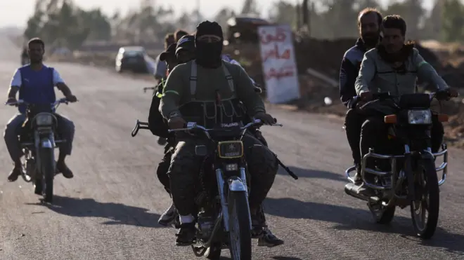 武装したベドウィンの戦闘員がシリア南部をバイクで移動している。手前2台のバイクには戦闘員が2人ずつ乗っている。戦闘員の1人は黒いフェイスカバーをつけている