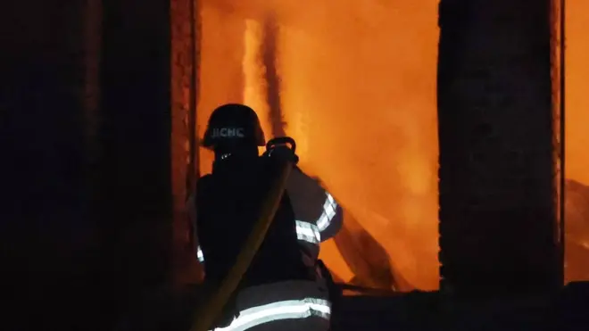 Пожежник стоїть спиною до камери, тримаючи в руках пожежний рукав. Він гасить пожежу.