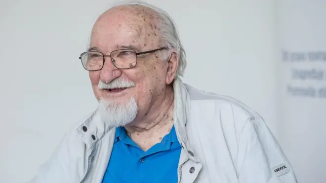 Sedi muškarac u sivoj jakni sedi i gleda u stranu, osmehuje se