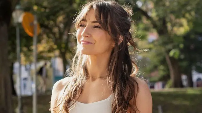 Sophie Macfie beyaz askılı tişörtüyle güneşli bir günde yukarı doğru bakıyor. Arkada yeşillikler var. 