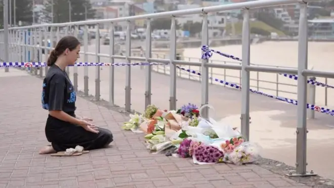 
砂浜を前にした遊歩道に手すりが並び、その前に多数の花束が供えられている。女性がその前に正座をして、両手を広げて手のひらを上に向け、目を閉じている。手すりには、白と青の制止線が張られている