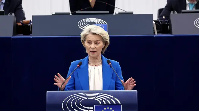 Mavi takım elbiseli sarışın bir kadın AB bayrağı olan bir kürsüde konuşuyor.