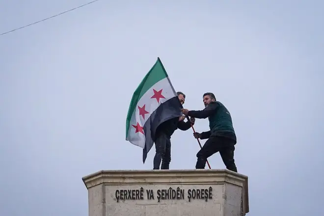 İki adam üç yıldızlı Suriye bayrağını anıta dikiyor.