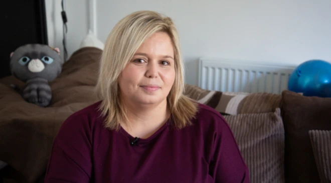 Danielle, mãe de quatro filhos, está sentada em uma poltrona na sala. Ela tem cabelos loiros e veste uma blusa cor de vinho. Ela olha para a câmera.