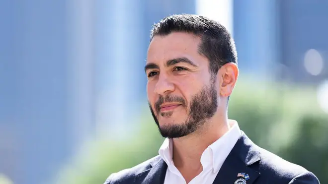 Sakallı kravatsız takım giyen genç sayılabilecek bir erkek mutlu görünüyor. Hava aydınlık ve güneşli