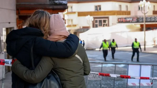 Две девушки в зимних куртках, одна в розовой шапке, вторая без головного убора, стоят, обнявшись за плечи, перед красно-белой полицейской лентой, огораживающей бар Le Constallation. За оцеплениием на заднем плане идут трое полицейских в ярких желто-зеленых жилетах