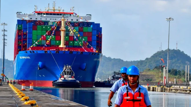 یک کشتی باری آبی رنگ در امتداد کانال پاناما حرکت میکند. دو کارگر با کلاه ایمنی آبی و جلیقههای نجات نارنجی در جلو ایستادهاند