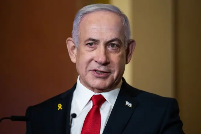 بنيامين نتنياهو مرتدياً بدلة رسمية سوداء عليها دبوس علم إسرائيل ودبوس آخر للتضامن مع الرهائن الإسرائيليين وربطة عنق حمراء