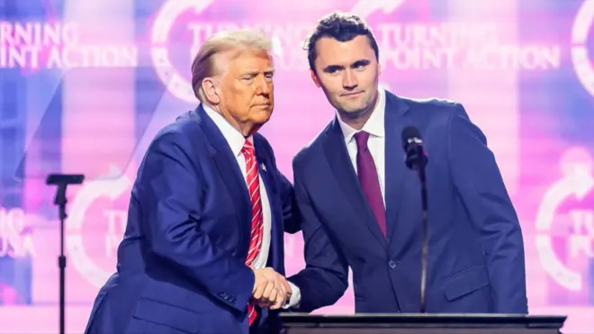 Donald Trump ściska dłoń Charliego Kirka na scenie. Oboje mają na sobie granatowe garnitury i czerwone krawaty.