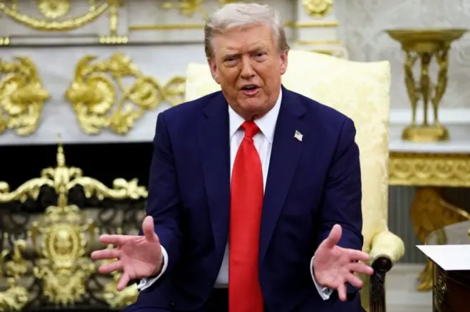 ترامب محركاً يديه ويتحدث. يرتدي بدلة رسمية كحلية اللون وقميصاً أبيض وربطة عنق حمراء، يجلس على كرسي يميل إلى اللون الذهبي في البيت الأبيض.