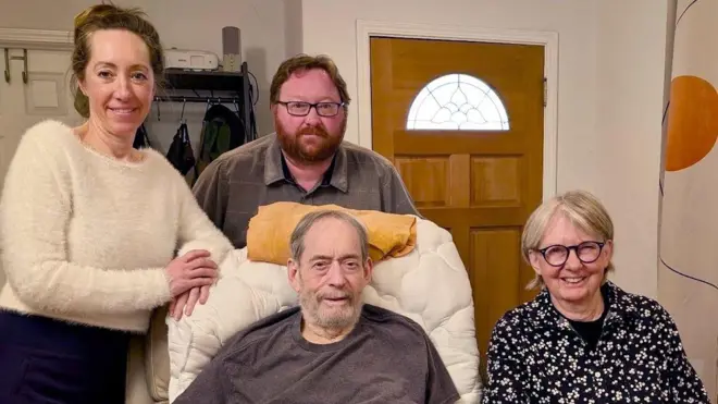 Un hombre se sienta en un sillón reclinable en pantalones cortos y camiseta en su sala de estar junto a su hija, su hijo y su esposa.