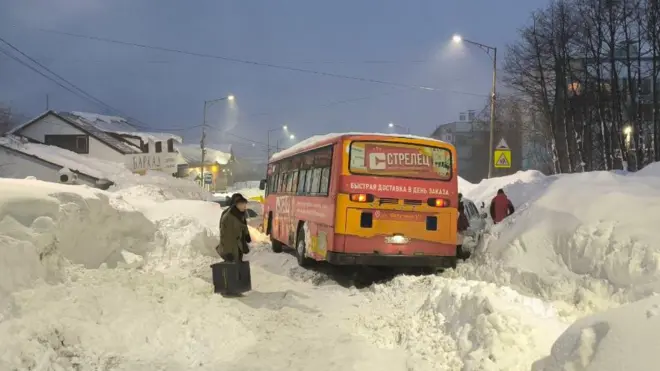 Автобус посреди снежных заносов, на фоне люди идут по неровным тропинкам.