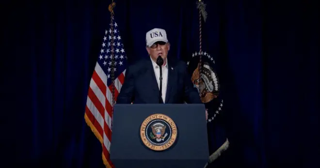 US President Donald Trump speaks at a podium with the US Seal while waring a dress shirt, dark blazer and white hat that is emblazoned with USA.