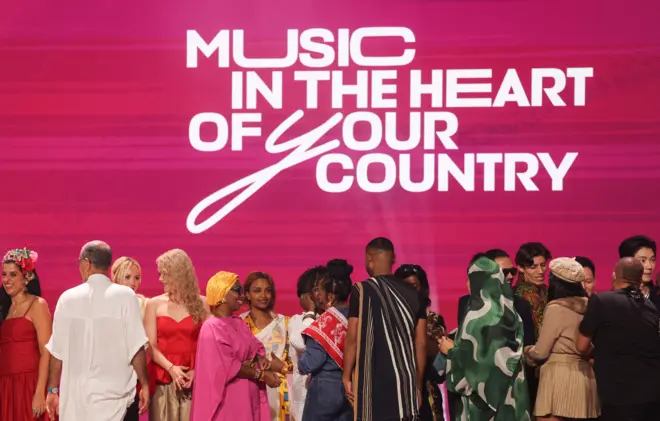 2025年9月12日，「Intervision」參賽者在莫斯科附近的俄羅斯國家中心參加抽籤儀式。現場人員身穿傳統服飾，在舞台上交流。舞台背景為亮粉紅色，白色字樣寫著：「音樂在你國家的心臟」。