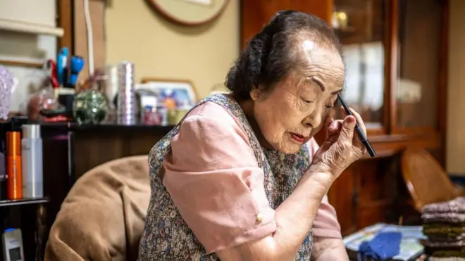 Tomoko Horino se dibuja las cejas con un lápiz de maquillaje mientras parece mirarse en un espejo. En el fondo, se observan otros implementos de belleza sobre encimeras.