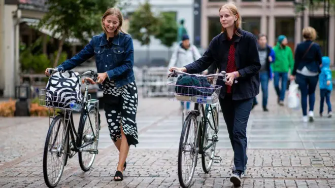 Dos mujeres caminan sonrientes empujando sendas bicicletas por una calle peatonal de Oslo. 