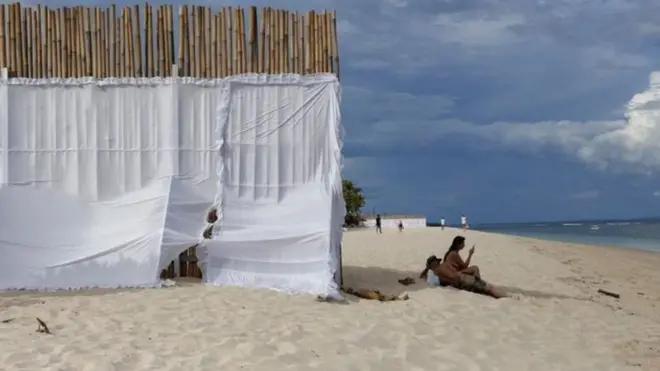 Dinding dari tongkat bambu dengan kain putih untuk menjaga privasi keluarga raja sudah didirikan di pantai dekat St Regis.