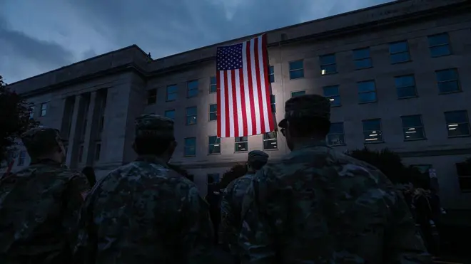 Група військових у камуфляжній формі, які стоять спиною до камери перед великою будівлею з кам’яним фасадом. На стіні будівлі висить великий прапор США, освітлений прожектором, що виділяє його на тлі сутінкового неба. Атмосфера урочиста, схожа на церемонію або вшанування.