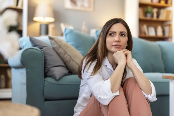 oturma odasında bir koltuğun önünde dizlerini karnında kavuşturarak yere oturmuş bir ellerini çenesine koymuş uzun kumral saçlı düşünceli bir kadın
