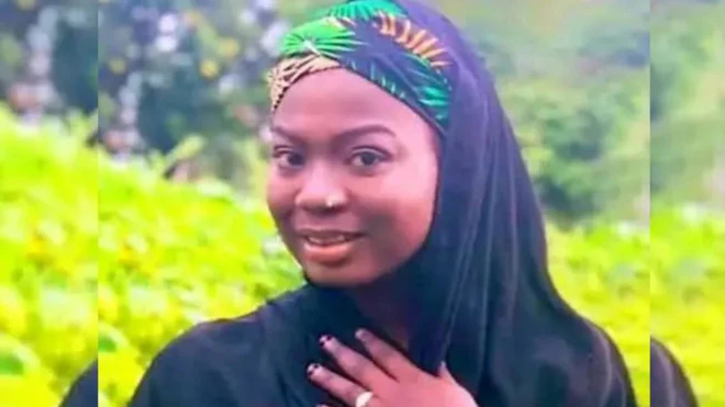 A lady looking at di camera wit brief smile, nose ring, hijab for her head and hand for her chest