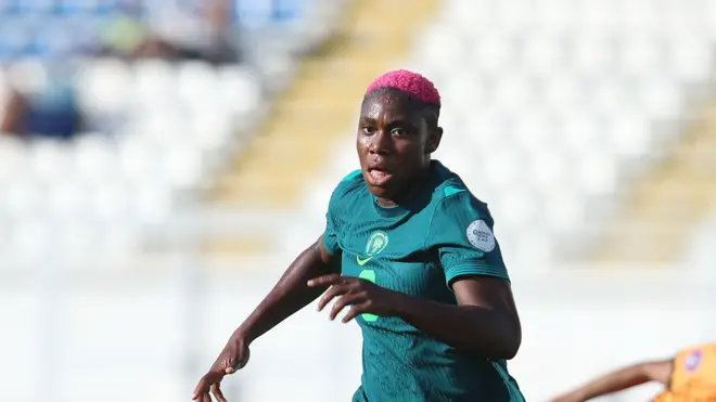 Asisat Oshoala, wearing an all-green Nigeria kit with number 8 on am, dey chase a ball during a match against Zambia, with three opponents in copper jerseys and socks and black shorts seen for di background on di green pitch. An empty stand with white seats dey blurred for di background
