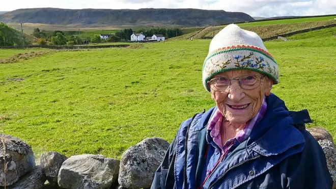 Margaret Bradshaw sonriendo a la cámara. Detrás de ella se ve un campo, una granja y colinas en el valle de Teesdale