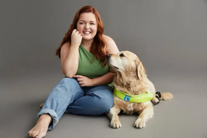 Lucy Edwards is sitting on the floor with her guide dog, Molly, to mark her becoming an RNIB ambassador. She has long red hair and is smiling. She is wearing a sleeveless green top and jeans 