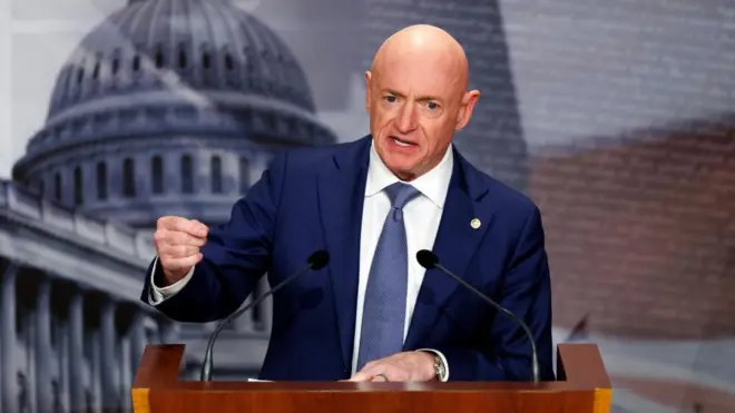 連邦議会の写真やアメリカ国旗のイメージを組み合わせた背景の前、マイクの載った演台に向かい、右手を動かしながら話す紺色のスーツ姿のケリー議員