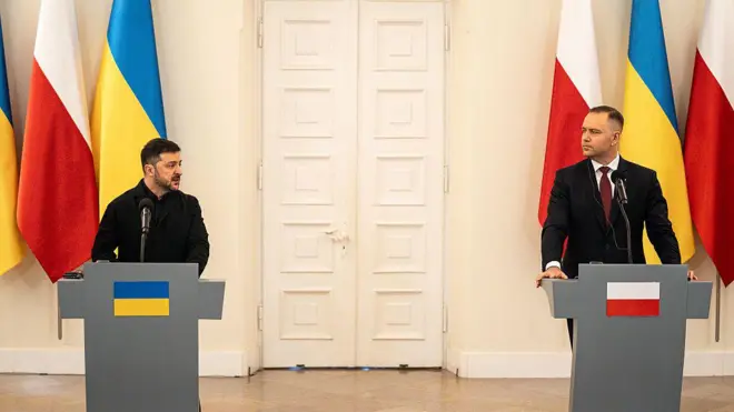 Серйозні Зеленський і Навроцький стоять за трибунами під час пресконференції на відстані кількох метрів. На задньому плані видно закриті білі двері та вертикально розташовані прапори Польщі та України.