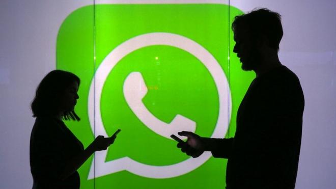 Dos personas sostienen sus celulares ante un cartel con el logotipo de WhatsApp.