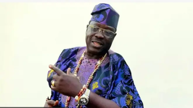 Taiwo Hassan Ogogo wọ Damaasi alawọ aro, o de fila, o lo ilẹkẹ sọrun o si fi aago sọwọ pẹlu ilẹkẹ. 