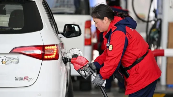 زنی با کاپشن قرمز رنگ در حال بنزین زدن به یک خودروی سفید