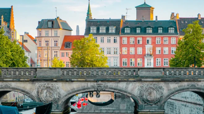 Zabytkowy kamienny most nad rzeką. W tle kamienice o czerwonych, jasnoróżowych i beżowych fasadach, rozłożyste drzewa i błękitne niebo. 