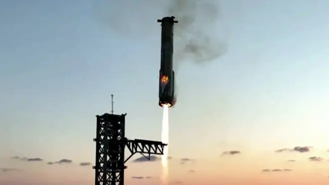The booster from the Starship rocket is shown landing between two mechanical arms with flames emitting from its base
