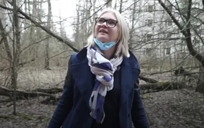 コートとマフラーを着用した女性が、森の中で上を見上げている。女性はめがねをかけ、あごにマスクをひっかけている