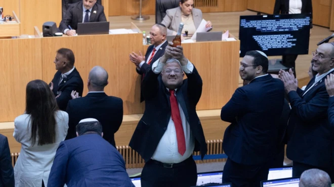 Bir takım elbiseli elinde kırmızı şampanya şişesi tutuyor. Yanındakiler ona gülüyor, bazıları da arlkışlıyor. Burası bir meclis salonu