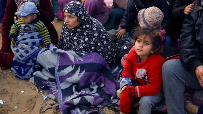 Les Palestiniens attendent d'être autorisés à retourner chez eux dans le nord de la bande de Gaza, après avoir été déplacés vers le sud sur ordre d'Israël pendant la guerre. Une femme portant un foulard est assise avec une couverture sur les genoux, avec un enfant portant un pull-over rouge à côté d'elle.