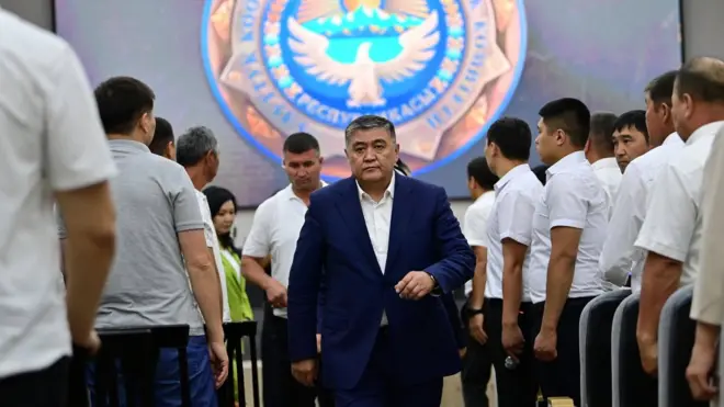 Оқ кўйлак ва тўқ кўк костюм-шим кийган Қамчибек Ташиев оқ кўйлакдаги эркак амалдорлар орасида юриб кетмоқда. 