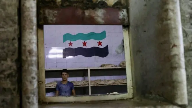عامل يقف في مخبز وخلفه علم (الثورة) السورية، بعد الإطاحة بالرئيس السوري بشار الأسد، في العاصمة السورية دمشق يوم 6 يناير/كانون الثاني 2025.