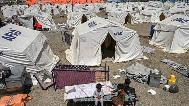  تصویر سه کودک در اردوگاه مهاجران در مرز پاکستان.این کودکان به روی تخت فلزی نشسته اند. اردوگاه خیمه‌های سفید با نشان یک شرکت خیریه دارد. 