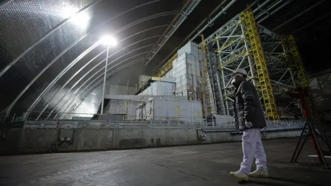 У великому закритому приміщенні з аркоподібною металевою стелею стоїть масивна промислова конструкція з бетону й металу, освітлена яскравими лампами. На передньому плані людина в захисному одязі, касці та масці повільно йде бетонною підлогою, поруч видно інженерне обладнання.