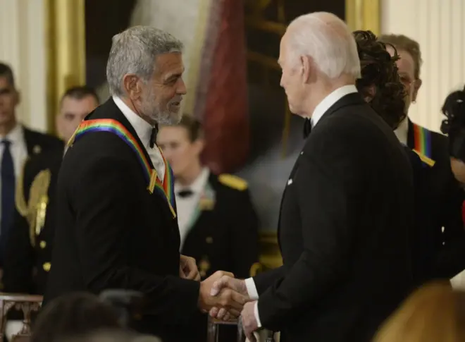 Biden e Clooney apertando as mãos em um evento. Ambos vestem ternos pretos
